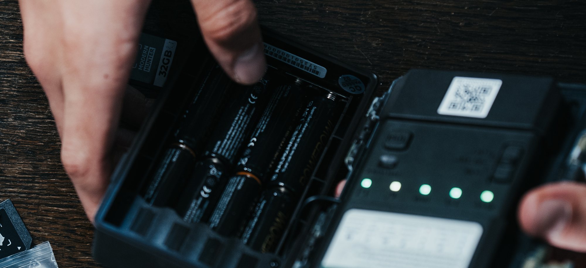 A set of AA cells being inserted into the battery compartment of a Modernhunter trail camera. Next to it lies a 32 GB memory card, and in the background a charger with green glowing charging bays.